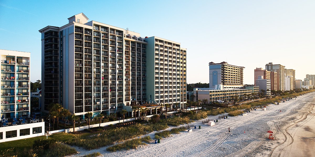 A large hotel building on a beach with sand and other buildings in the background