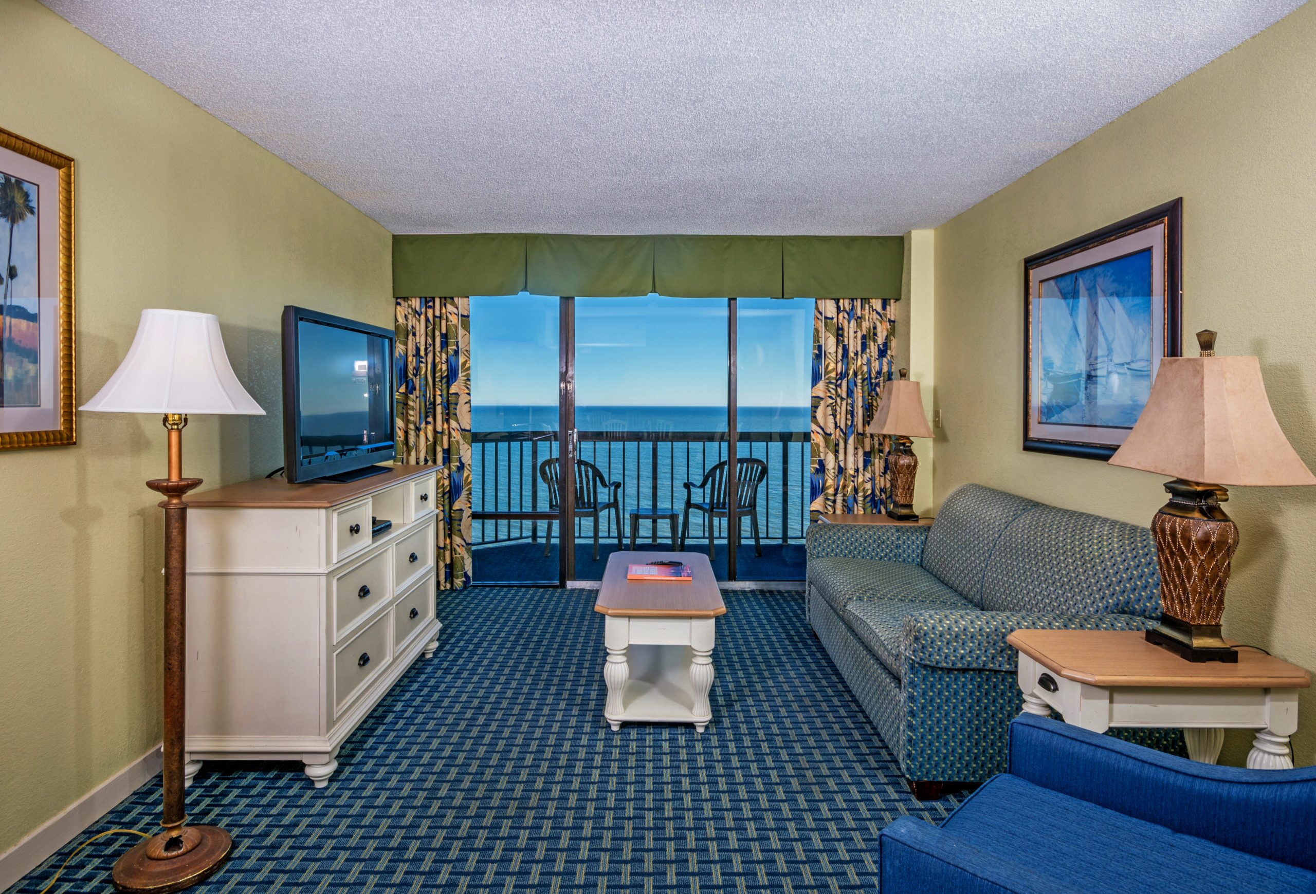 A wide view of a hotel room's living area with a sofa, TV, coffee table, and balcony with ocean view.